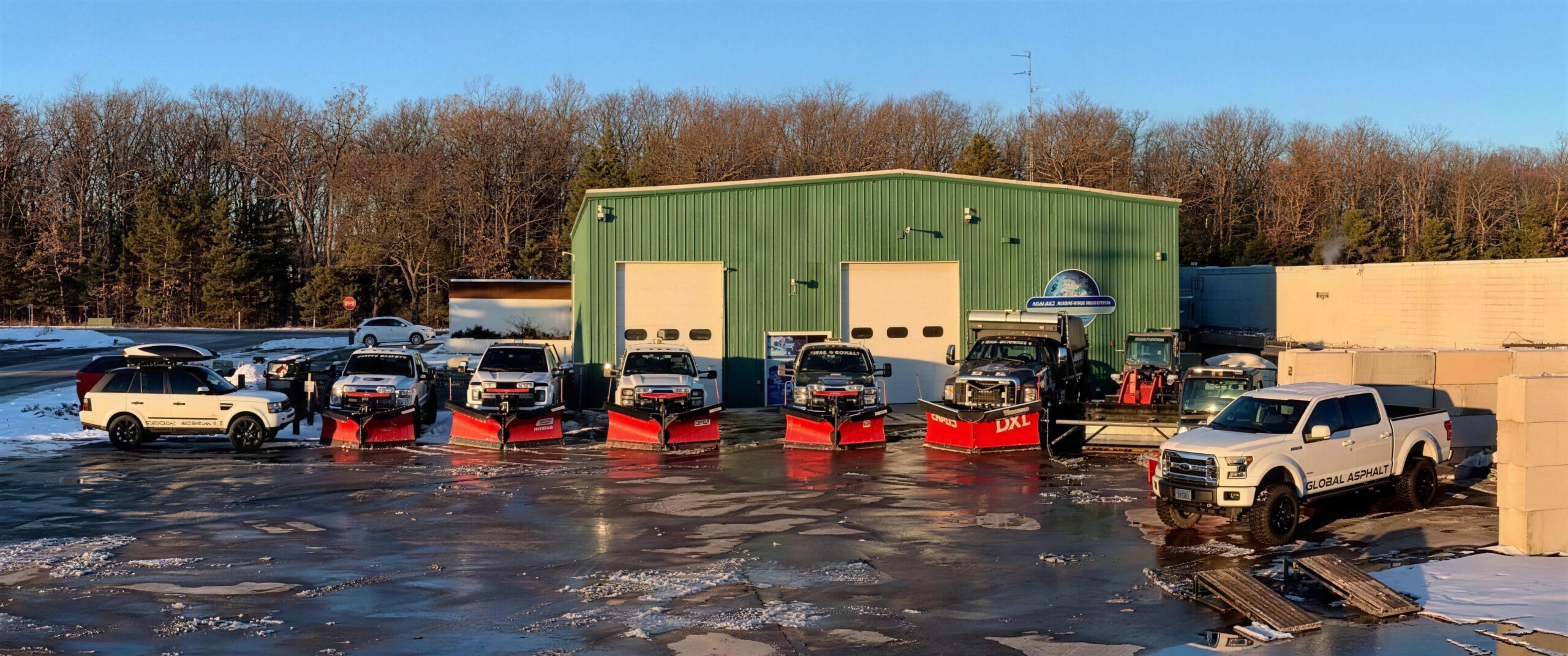 Global Asphalt plow trucks lined up outside the shop at dusk, blades down, ready for the night