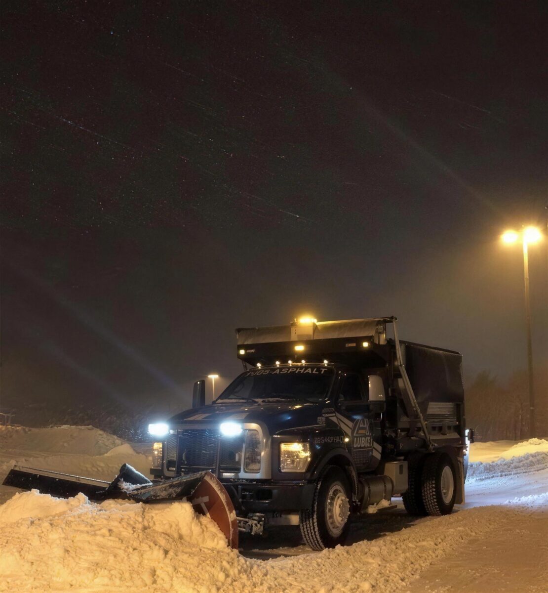 Global Asphalt plow truck clearing a commercial lot at night in a snowstorm